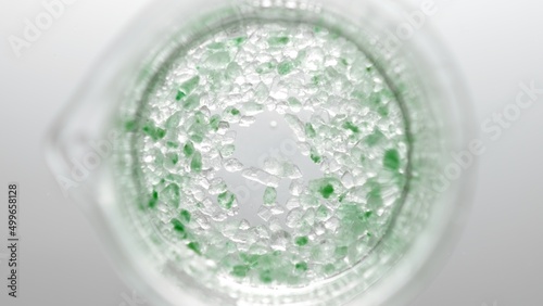 Top view shot of beaker with crystals of green salt in water on light grey background | Mineral skin care cosmetics formulation concept
