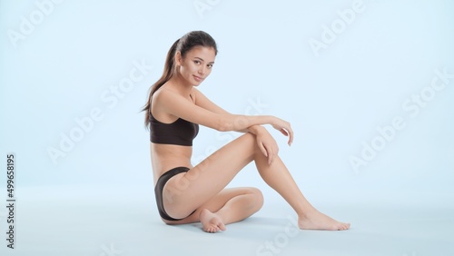 Young slim Asian woman with long dark hair in ponytail in black bikini sits on the floor and looks at camera on pale blue background | Body care concept