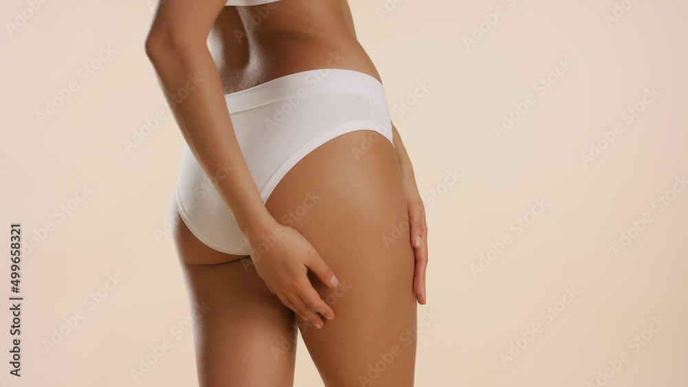 Horizontal close-up of slim good-looking woman in white underwear strokes her hip on beige background | Anti cellulite concept