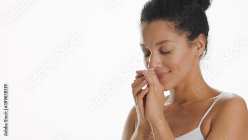 Young African American woman in white top smells her hands enjoying the fragrance on white background | Hand care concept