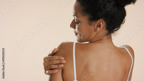 Horizontal close-up of beautiful slim African American woman with curly hair strokes her shoulder standint with her back to camera on beige background | Skin nourishing concept photo