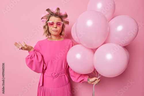 Crying displeased woman with leaked makeup wears shades and dress stands upset has spoiled party makes hairstyle holds bunch of inflated balloons isolated over pink background. Monochrome shot