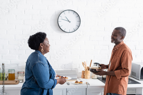 happy senior african american couple looking at each other while holding pancakes and coffee pot in kitchen. photo