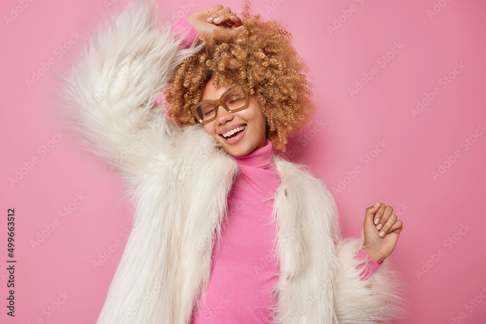 Carefree optimistic attractive woman with curly hair shakes arms dances to music wears casual turtleneck and white fur coat smiles joyfully moves against pink studio wall being full of happiness. Carefree optimistic attractive woman with curly hair shakes arms dances to music wears casual turtleneck and white fur coat smiles joyfully moves against pink studio wall being full of happiness.