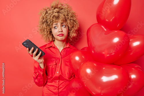 Displeased young European woman has spoiled mood waits forlover on Valentines Day uses mobile phone feels lonely holds bunch of heart balloons has leaked makeup isolated over red background.