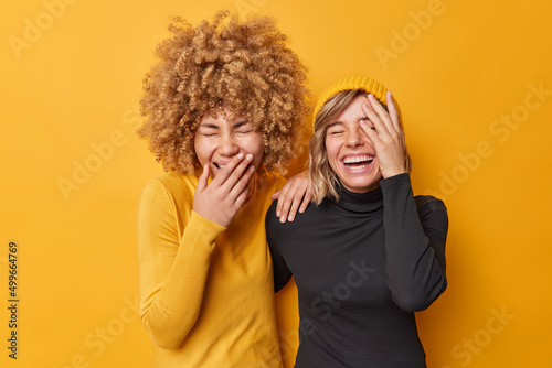 Photo of overjoyed friendly women laugh out loudly giggle as hear funny joke have fun chuckle at camera feel positive dressed in casual turtlenecks isolated over yellow background. Happiness concept photo