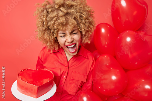 Photo of emotional displeased woman with curly hair holds sweet cake prepared for holiday bunch of helium balloons celebrates birthday comes on party isolated over red background. Celebration