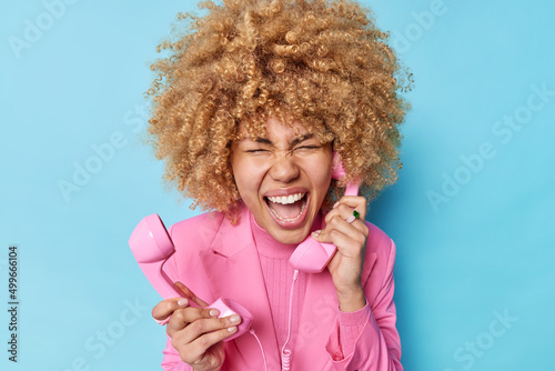 Angry emotional woman holds two telephone receivers screams loudly in retro phone handset irritated by customer call wears pink formal jacket poses over blue background complains on connection quality