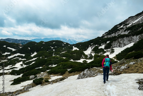 Woman with backpack crossing a snow field on a hiking trail with view on cloud covered mountain peaks of the Hochschwab Region in Upper Styria, Austria. Plateau full of snow in beautiful Alps, Europe.