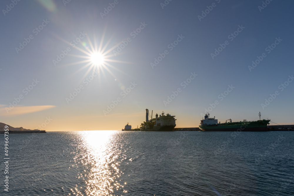 Large ships moored in the harbor in Motril with the sun shining in the sky reflecting on the water, Andalucia Spain.