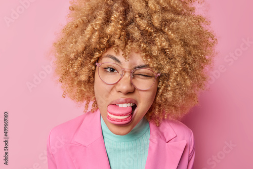 Headshot of pretty woman with curly bushy hair holds delicious yummy doughnut in mouth winks eye wears transparent eyeglasses and formal jacket isolated over pink background. Sugar diet or addiction