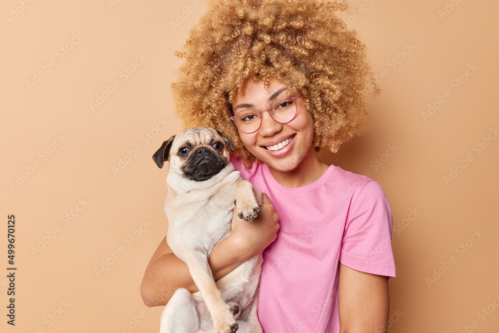 Portrait of cheerful young woman hugs her favorite pug dog smiles gladfully expresses love and care to pet isolated over brown background. Veterinary health concept. Owner and pet relationships Portrait of cheerful young woman hugs her favorite pug dog smiles gladfully expresses love and care to pet isolated over brown background. Veterinary health concept. Owner and pet relationships