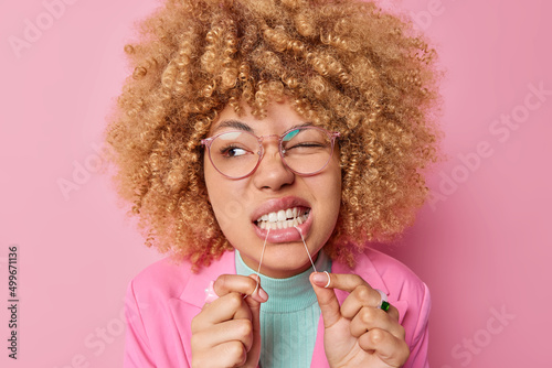 Dental health care concept. Beautiful young woman with curly bushy hair cleans teeth with dental floss wears transparent eyesglasses isolated over pink background shows how to floss correctly photo