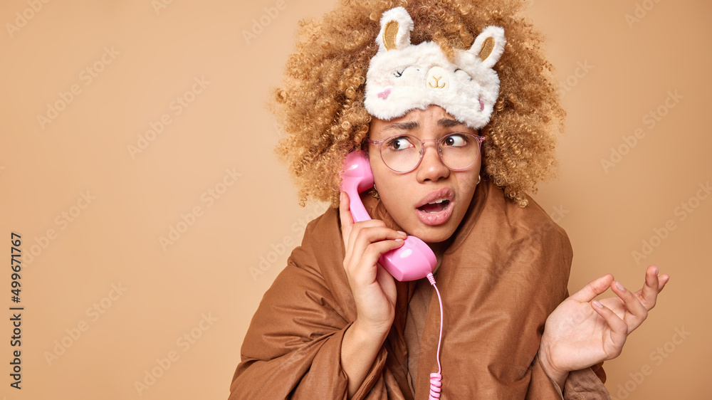 Hesitant woman feels displeased looks away confused shrugs shoulders wears blindfold wrapped in blanket talks via handset feels unaware poses against brown background copy space for your advertising