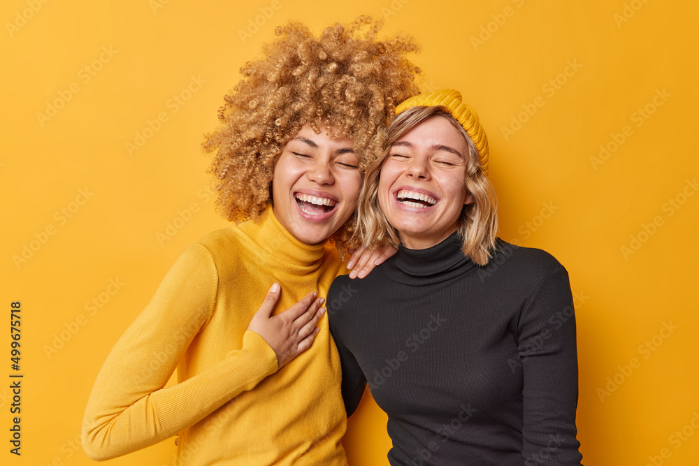 Sincere human emotions concept. Positive overjoyed young women have fun laugh gladfully smile toothily cannot stop laughing stand closely to each other dressed casually isolated over yellow wall - obrazy, fototapety, plakaty 