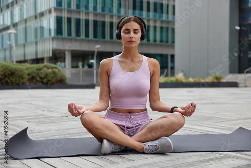 Relaxed calm woman sits in lotus pose on fitness mat meditates and listens pleasant music via headphones practices yoga in urban setting wears sportsclothes keeps eyes closed breathes deeply