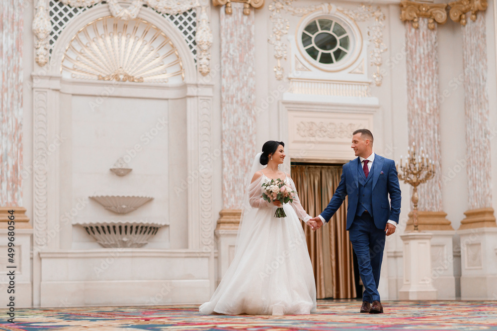 Front view o cute and pretty girl in long wedding dress, holding flowers arrangement, smiling and looking at bridegroom while walking with him inside of luxury hall