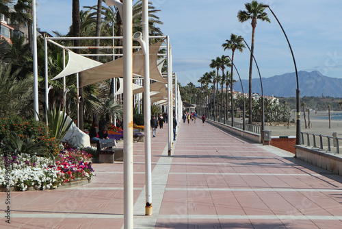 ESTEPONA, SPAIN - MARCH 3RD 2020: People stroll along the esplanade. Canvas sails provide shade in the heat of the day