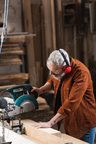 tattooed carpenter cutting wood with miter saw in woodwork studio.