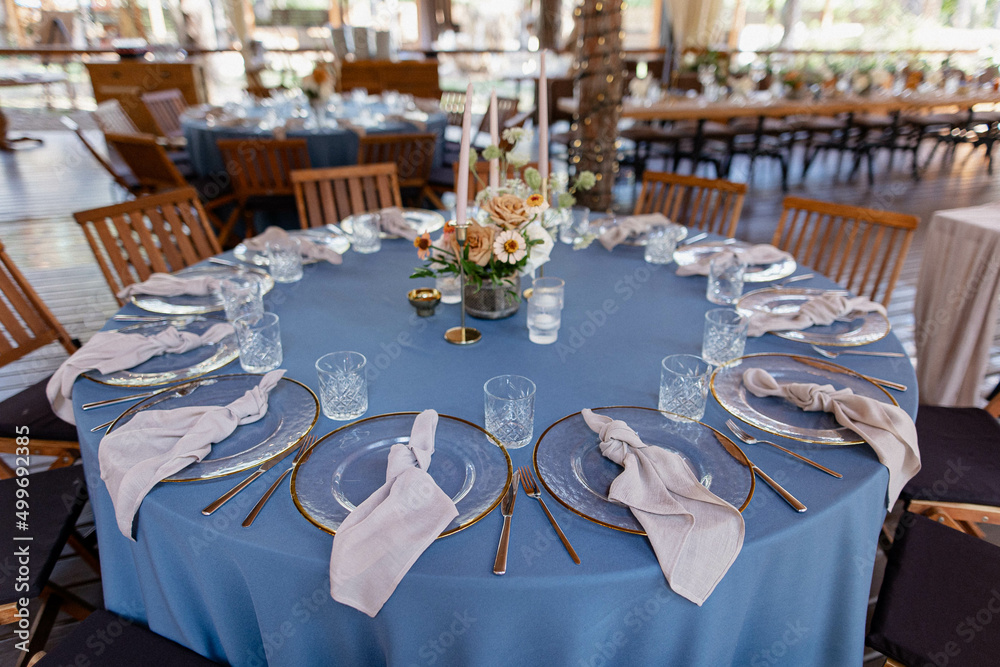 Wedding Decor. Festive table decorated with flowers on the center, candles, silverware and plates with silk napkins on dusty blue tablecloth