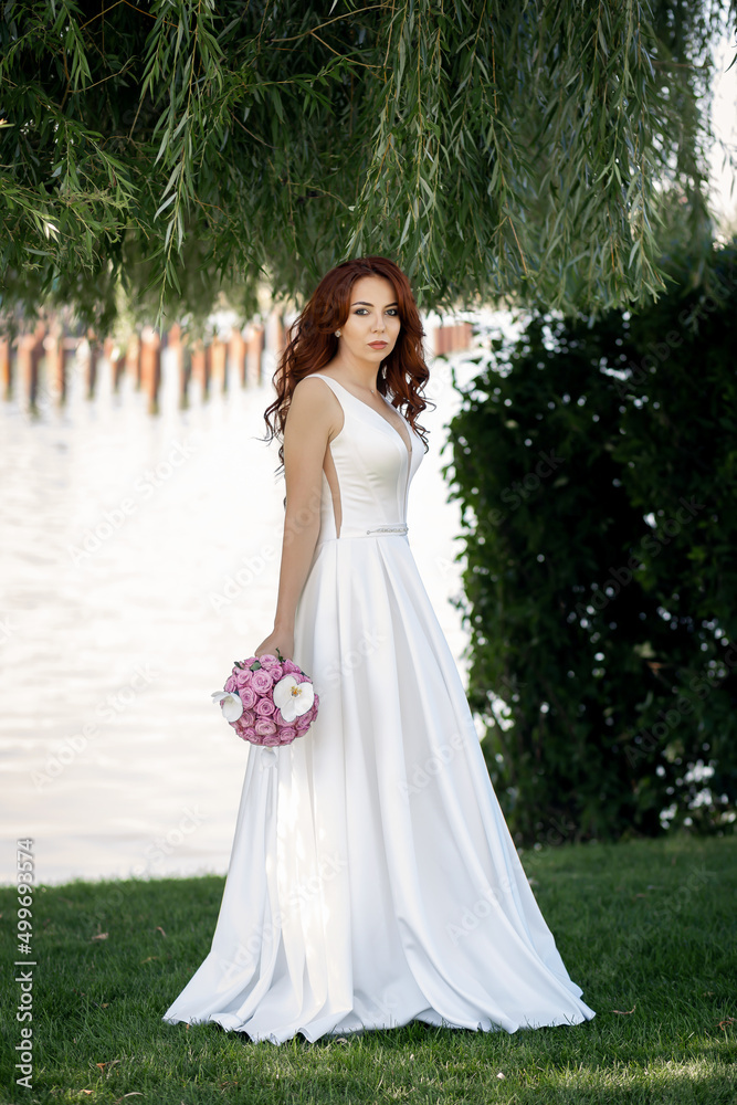 Refined feminine lady with red hair in a white dress. A happy and satisfied bride poses in a green park. Summer mood