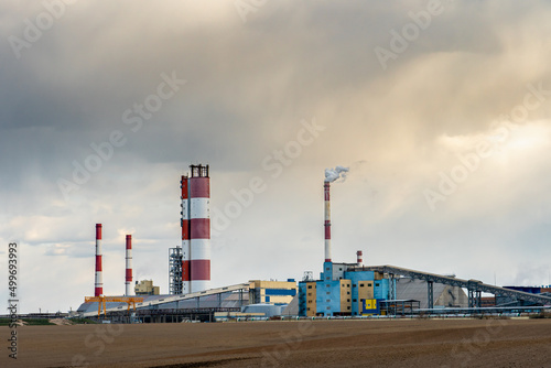 A working chemical industry plant against the background of clouds and an agricultural field. Factory pipes emit columns of smoke poisoning the environment. Poisoning of agricultural land with waste