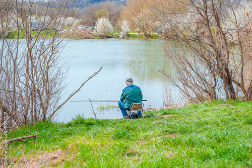 Rear view of an elderly gray-haired man fishing on a lake in nature, patience and relaxation concept. Portrait of a senior fisherman with a fishing rod sitting on a chair in nature, in the countryside