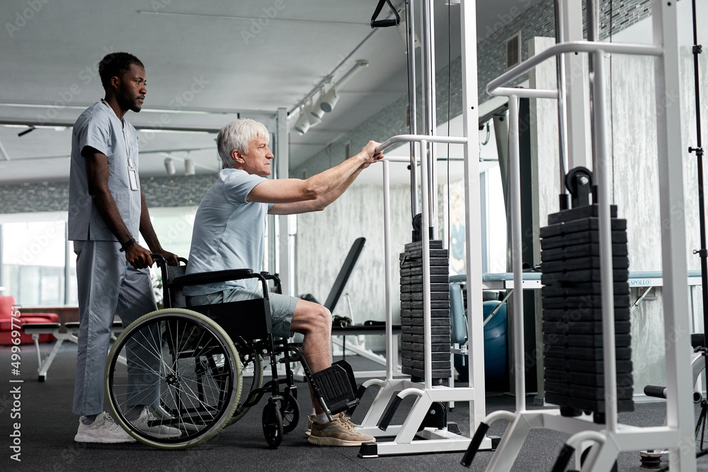 Side view portrait of senior man in wheelchair using exercise machines at rehabilitation clinic, copy space Side view portrait of senior man in wheelchair using exercise machines at rehabilitation clinic, copy space