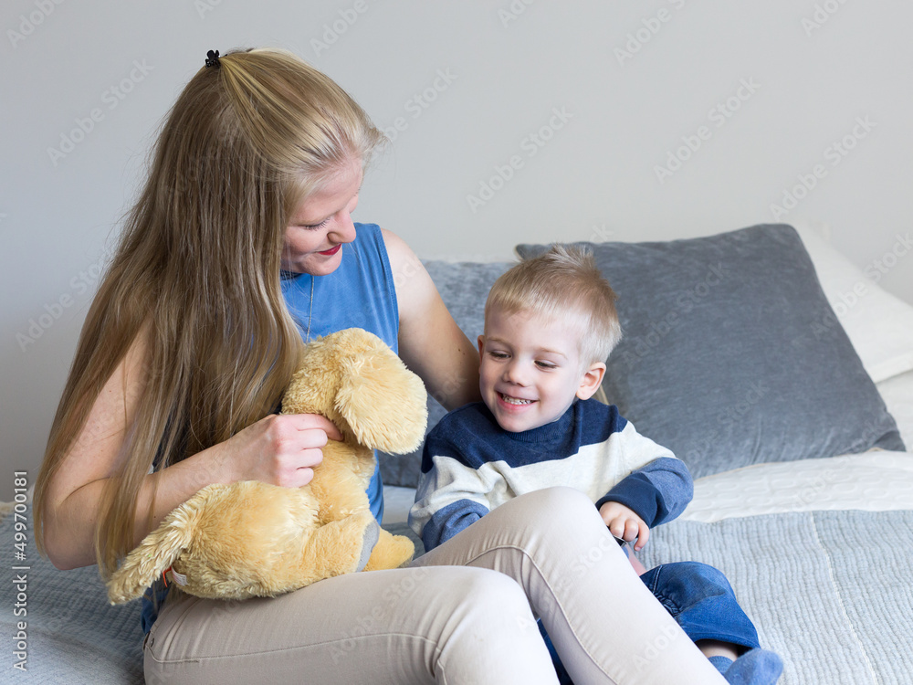Cute three-year old blond boy sitting on bed dressed in casual clothes playing with his mother and a stuffed toy Cute three-year old blond boy sitting on bed dressed in casual clothes playing with his mother and a stuffed toy
