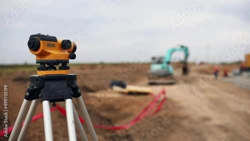 Optical level mounted on tripod against machinery operating at rural site. Excavator digs trench for laying power cables on cloudy day close view photo