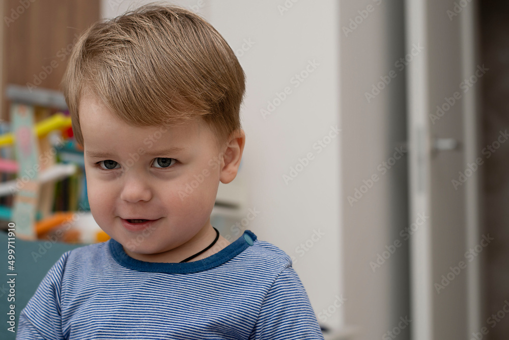 Little caucasian handsome boy emotionally frowns his eyebrows and smiles in a home interior in a striped sweater. Little caucasian handsome boy emotionally frowns his eyebrows and smiles in a home interior in a striped sweater.