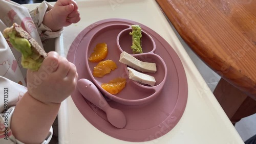 Hands of baby holding avocado bread stick sitting on high chair with tofu and orange pieces on rubber plate. Baby led weaning, healthy food concepts photo