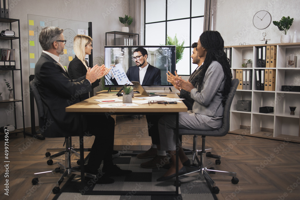 Group of four skillful multiethnical businesspeople clapping hands and looking at screen, having video conference in boardroom, with their male colleague explaining some financial charts. Group of four skillful multiethnical businesspeople clapping hands and looking at screen, having video conference in boardroom, with their male colleague explaining some financial charts.