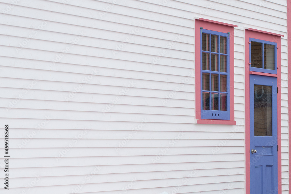 Fototapeta premium The exterior wall of a white wooden cape cod clapboard siding house with a purple panel door and vintage glass window, black metal hinges, pink trim around the door, and white snow on the step.