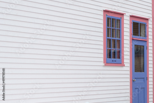 The exterior wall of a white wooden cape cod clapboard siding house with a purple panel door and vintage glass window, black metal hinges, pink trim around the door, and white snow on the step.  © Dolores  Harvey