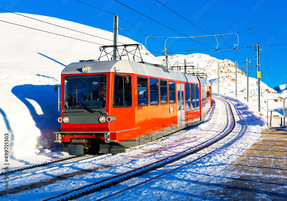 Modern red train running on rack railway through snowy Swiss Alps on sunny winter day.