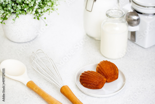 Madeleine cookie or French sponge cake in a white plate alongside white kitchen utensils, sugar, milk and flour jars on white background.