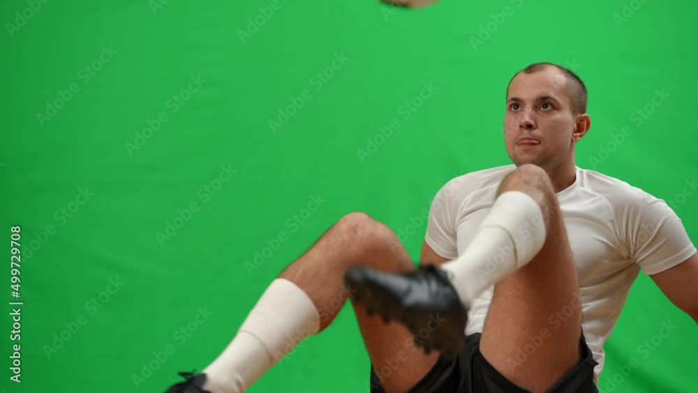 Professional confident footballer juggling catching ball with legs sitting on green screen and looking away smiling. Positive Caucasian sportsman exercising working out at chromakey background Professional confident footballer juggling catching ball with legs sitting on green screen and looking away smiling. Positive Caucasian sportsman exercising working out at chromakey background