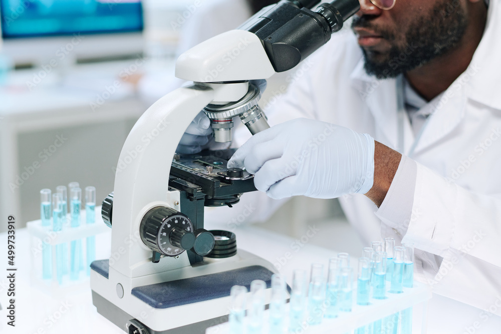 Gloved hands of modern African American male scientist studying new virus in microscope while sitting by workplace with test tubes Gloved hands of modern African American male scientist studying new virus in microscope while sitting by workplace with test tubes
