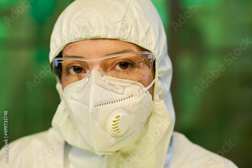 Face of young female scientist in protective eyeglasses, respirator and coveralls standing in front of camera in clinical laboratory photo