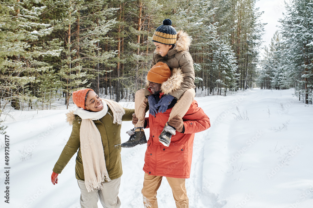 © pressmaster - Young contemporary family of father, mother and son enjoying winter day in park while African American man carrying little boy on shoulders