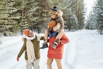 Young contemporary family of father, mother and son enjoying winter day in park while African American man carrying little boy on shoulders
