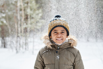 Cheerful little boy of African ethnicity wearing warm winter jacket and beanie hat while having fun in the forest on frosty day