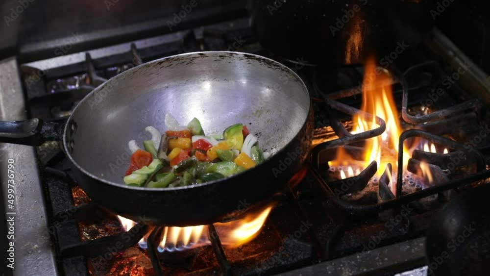 Culinary. Asian cuisine. Closeup view of a chef cooking sliced vegetables in the wok pan. The food catches fire and the chef shakes the wok. Culinary. Asian cuisine. Closeup view of a chef cooking sliced vegetables in the wok pan. The food catches fire and the chef shakes the wok.