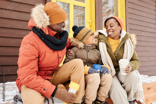 Cheerful black woman in winterwear looking at her cute son during talk while sitting on porch of cottage next to young man and having tea