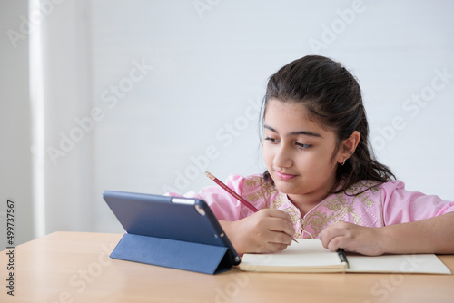 cute Indian girl writing on notebook and looking tablet for online learning class, education concept