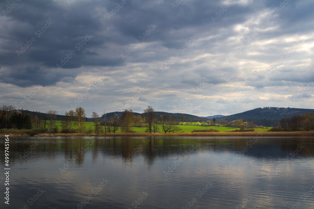 Small pond with spring grass and tress on shore under dramatic day sky. Czech landscape