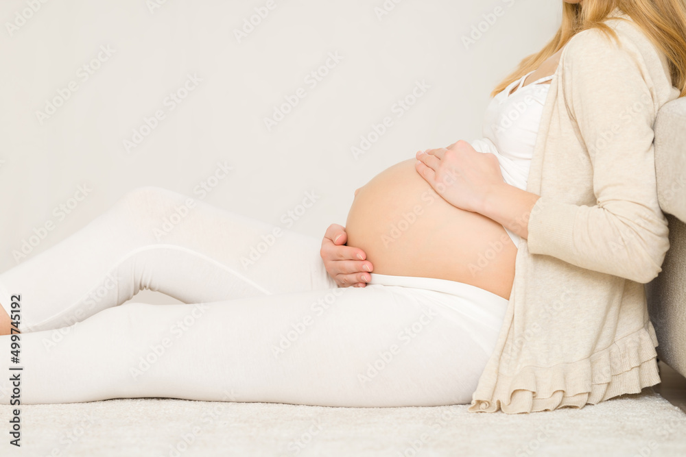 Young adult pregnant woman sitting on carpet at living room, touching big naked belly and feeling baby move. Pregnancy concept. Expectation time. Closeup. Side view. Young adult pregnant woman sitting on carpet at living room, touching big naked belly and feeling baby move. Pregnancy concept. Expectation time. Closeup. Side view.