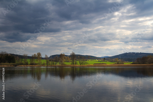 Small pond with spring grass and tress on shore under dramatic day sky. Czech landscape