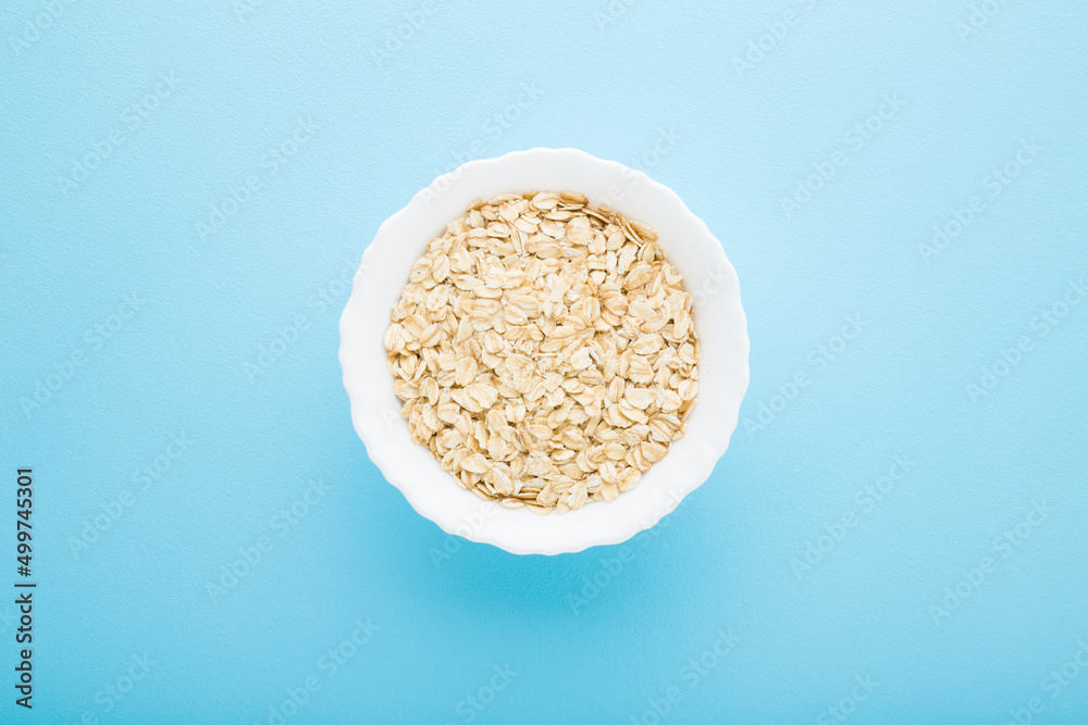 White bowl with dry rolled oat on light blue table background. Pastel color. Closeup. Healthy food. Top down view.
